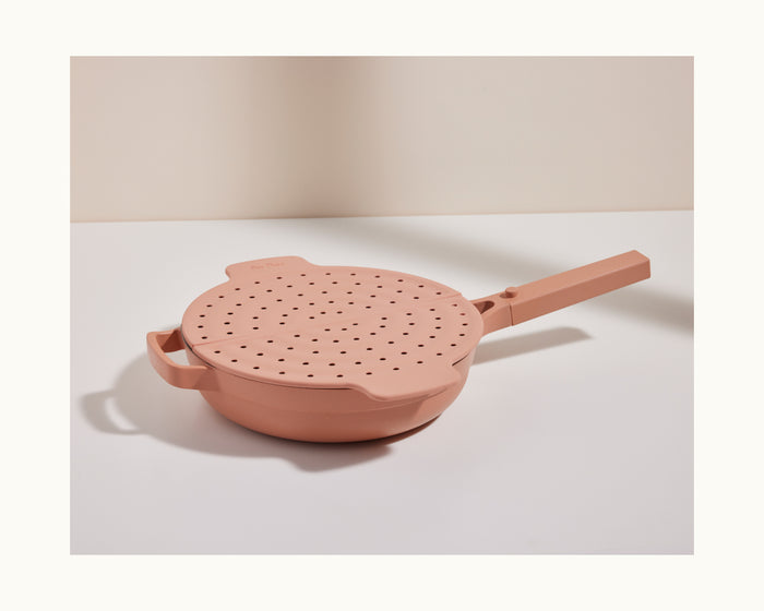 The Fearless Fry, a pink nonstick frying pan inspired by the Always Pan, comes with a matching handled lid featuring multiple steam holes for splatter prevention and sits on a white surface against a neutral background.