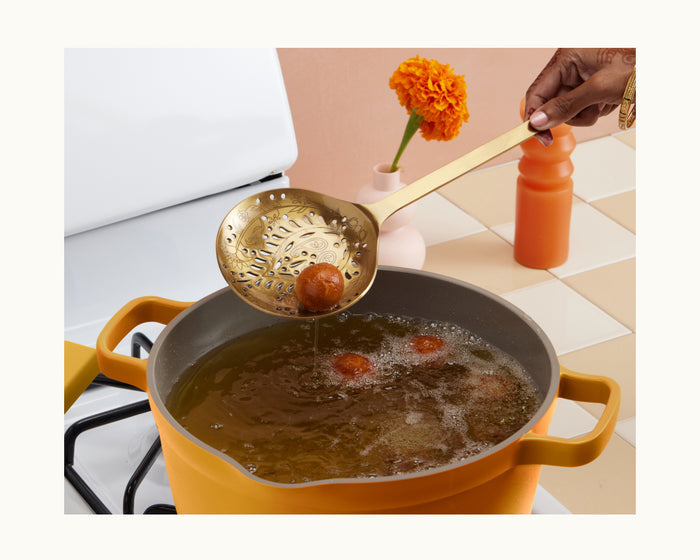 Using the Golden Fry Set, a person lifts a fried dessert ball from hot oil in a yellow pot, with marigold flowers and a bottle nearby, evoking the festive spirit of Diwali.