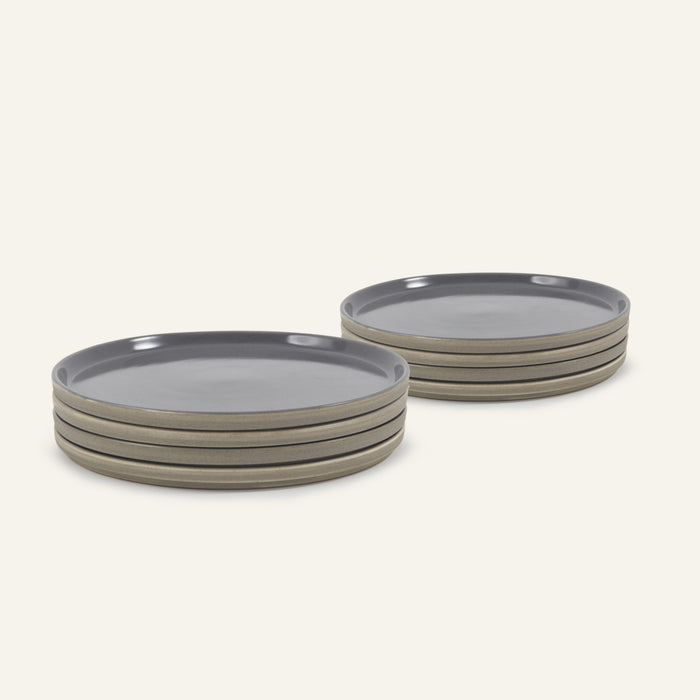 Two stacks of Main Plates, round and hand-painted gray with beige sides, are arranged on a light background. Each stack has four plates, highlighting a modern minimalist style that’s both functional and stylish.
