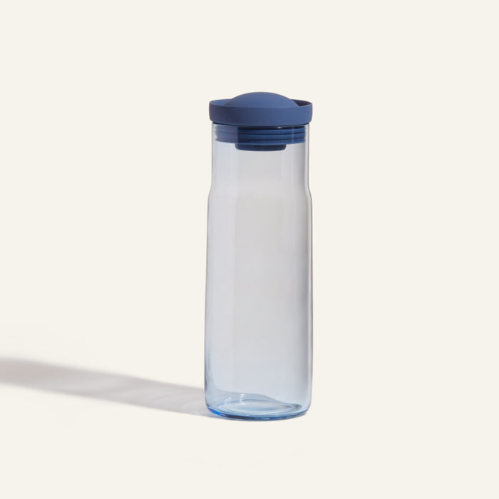 The Night + Day Carafe is a transparent plastic water bottle with a blue screw-on lid, standing upright on a plain light background and featuring a sleek, modern carafe design.