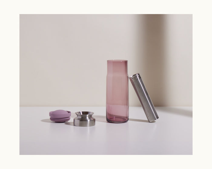 The Night + Day Carafe, a tall translucent purple glass piece, stands beside a stainless steel cylinder, a small metal funnel, and two round purple caps on a white surface.