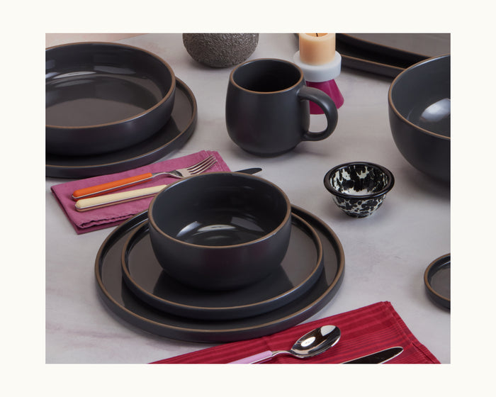 A table set with hand-glazed ceramic salad plates, bowls, and a mug alongside silverware on red napkins, a small patterned bowl, and candles in the background.
