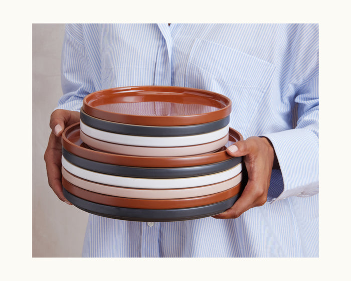 A person wearing a blue and white striped shirt holds a stack of Salad Plates—round, hand-glazed ceramic dishes with brown, white, and black bands—perfect for adding charm to any tableware collection.