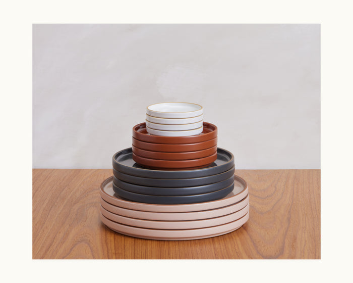 A stack of Salad Plates in various sizes and colors—white, terracotta, gray, and beige—arranged on a wooden surface against a light background, perfect for displaying your favorite tableware.