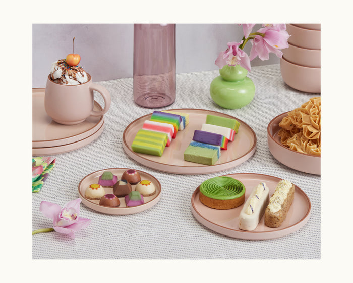 A table set with hand-glazed Salad Plates in pastel pink holds assorted colorful desserts, kueh, pastries, cookies, and a mug of whipped cream with a cherry, with vases of flowers in the background.
