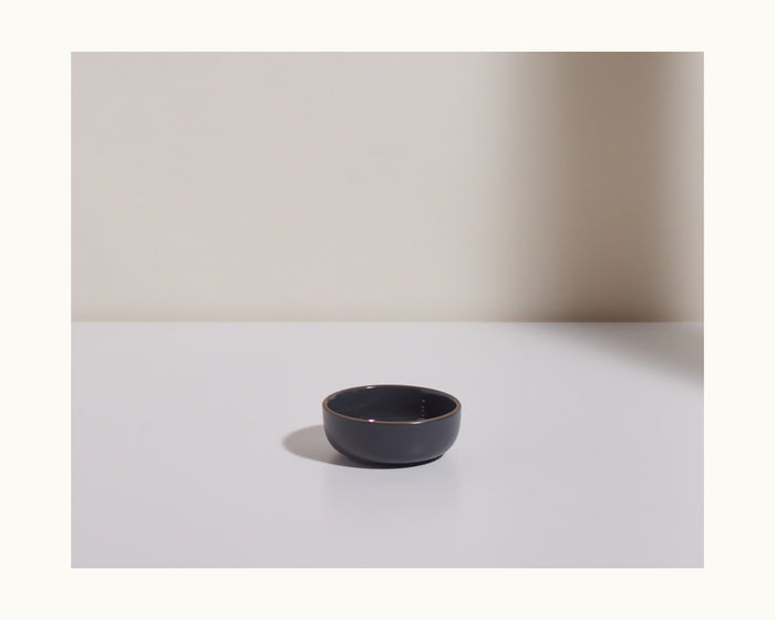 A Tiny Bowls black ceramic piece sits on a white surface with a right-side shadow against a neutral background—ideal for modern kitchens wanting stylish, stackable tableware.