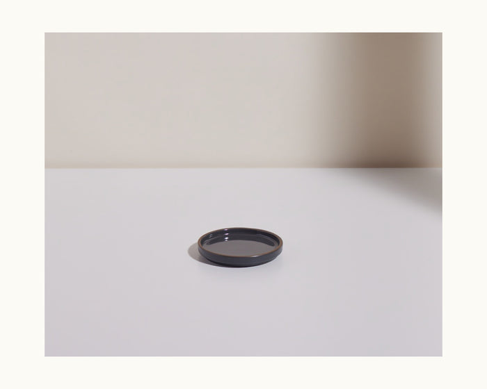 The Tiny Plates product is a small, round black ceramic plate placed on a white surface with a light background, casting a shadow to the right.
