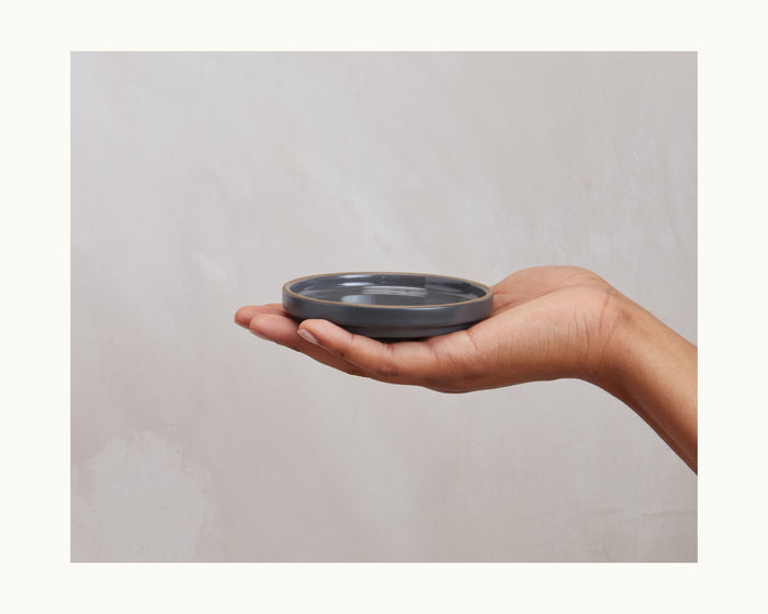 A hand displays the Tiny Plates, a flat, round ceramic plate, against a plain light-colored background.