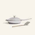The Titanium always pan pro non toxic frying pan, a white lidded frying pan with a long handle and innovative nonstick technology, sits next to a metal spatula with a beige handle on a light background.