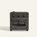 The AU - Wonder Oven is a dark gray countertop toaster oven with three top control dials and a front glass door featuring a diamond-patterned interior. "Our Place" appears at the bottom of the door. Doubles as an air fryer for extra versatility.