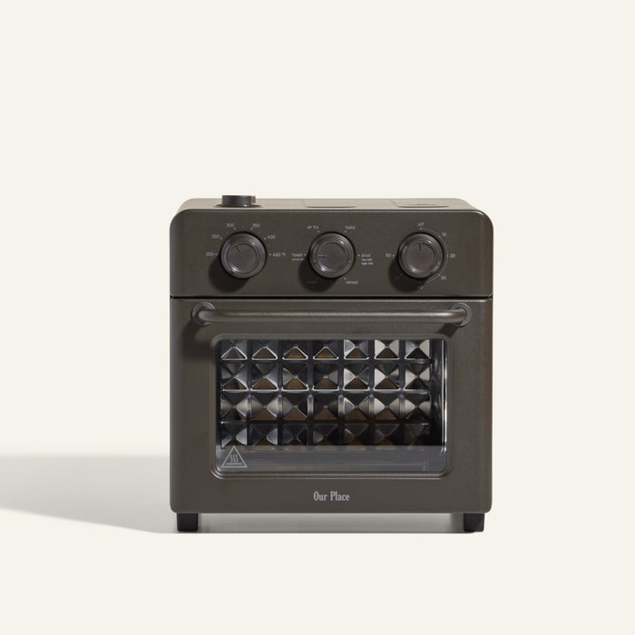 The AU - Wonder Oven is a dark gray countertop toaster oven with three top control dials and a front glass door featuring a diamond-patterned interior. "Our Place" appears at the bottom of the door. Doubles as an air fryer for extra versatility.